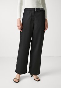 Zwarte wijde broek met een platte voorkant, voorzien van een riem met een metalen gesp. Glad materiaal en scherpe plooien langs de benen.