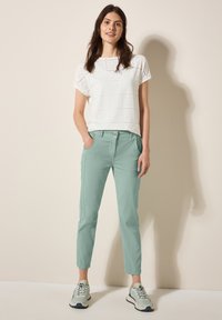Weißes Kurzarmoberteil mit Ausschnitten, kombiniert mit hellmintgrünen Hosen. Das Model trägt graue und beige Sneaker. Einfacher Hintergrund.