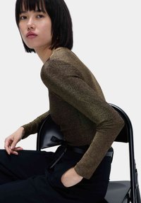 Top de manga larga metálico en bronce con un brillo sutil, combinado con pantalones negros. Modelo sentado en una silla negra, mostrando una pose relajada.