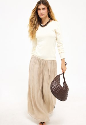 Žena s dlhými vlasmi, ktorá nosí krémový sveter a béžovú plisovanú sukňu, drží v pravej ruke hnedú pletenú kabelku.