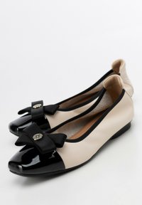 Zapatos planos negros y beige con punta de charol, detalle de lazo en la parte frontal y un tacón bajo y sólido. Textura de cuero suave.