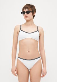 Hvit bikinisett med tynne svarte stropper og merkevare langs kantene. Glatt tekstur og minimalistisk design. Inkluderer matchende bikinitruser.