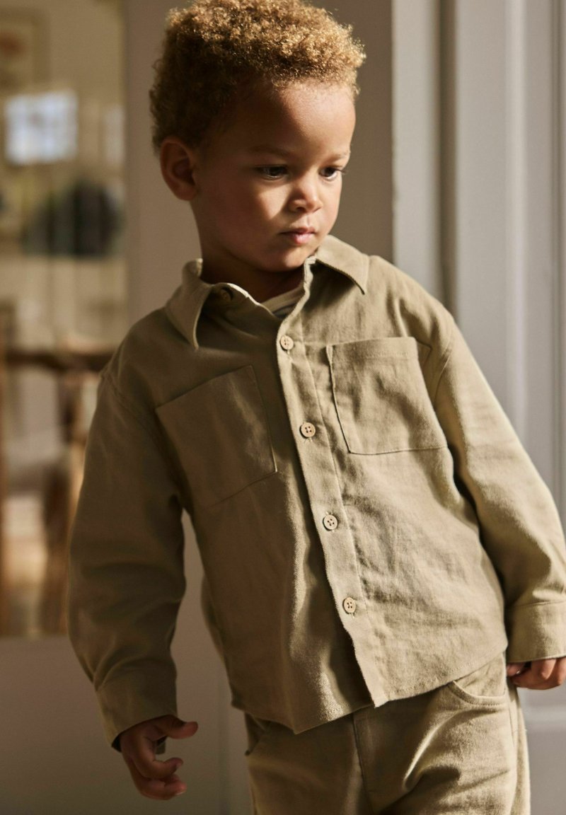 Jeune enfant aux cheveux bouclés portant une chemise beige boutonnée et un pantalon assorti, debout à l'intérieur près d'une fenêtre avec une lumière douce sur le visage.