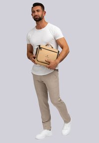 Bolso de mano sintético beige con detalles y cremalleras negras, que cuenta con un asa superior. El modelo lleva una camiseta blanca y pantalones beige claros.
