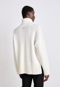 Fetter weißer Pullover mit hohem geripptem Kragen, vertikaler Rippenstruktur auf der Rückseite und lockerer Passform; akzentuiert durch ein dezentes Markenlogo.