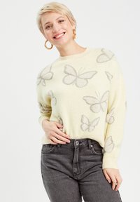 Lys gul sweater med grå sommerfuglemønstre, rund halsudskæring, ribbede manchetter og kant, parret med mørkegrå jeans.