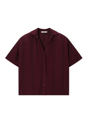 Femme portant des lunettes de soleil écaille, une chemise bordeaux ample boutonnée avec broderie, un short assorti, tenant un sac tressé sur un fond beige.