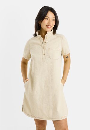 Femme souriante portant une robe texturée crème à manches courtes avec poche avant et col boutonné, mains dans les poches, sur fond blanc uni.