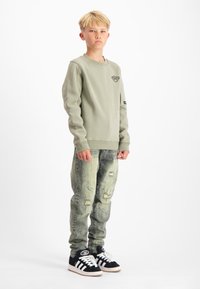 Groene sweatshirt met een klein logo, gecombineerd met verouderde lichtblauwe jeans en zwarte sneakers met witte strepen en logo.