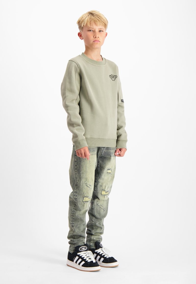 Groene sweatshirt met een klein logo, gecombineerd met verouderde lichtblauwe jeans en zwarte sneakers met witte strepen en logo.