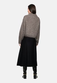 Pull en maille épaisse marron avec un col haut, associé à une jupe noire fluide. Le modèle porte des bottes noires brillantes jusqu'au genou.