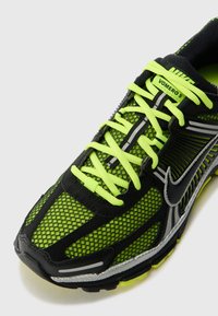 Zapatilla deportiva negra y verde neón con parte superior de malla, acentos reflectantes y cordones brillantes, que cuenta con una suela acolchada y detalles de diseño intrincados.