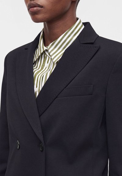 Gros plan d'une personne portant un blazer noir croisé sur une chemise à rayures verticales vertes et blanches.