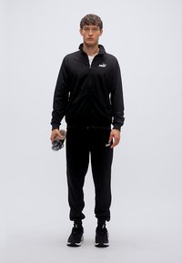 Μαύρη φόρμα με τζάκετ και παντελόνι με φερμουάρ και διακοσμητικά λογότυπα. Το μοντέλο κρατάει ένα παγούρι νερού και φοράει μαύρα αθλητικά παπούτσια.