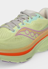 Chaussure de course vert clair avec lacets beige, logo violet et bande orange sur la semelle, montrant une tige texturée et respirante et une semelle intermédiaire rembourrée.