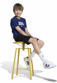 Geel metalen kruk met een geometrisch ontwerp. Kind met een blauwe shirt, zwarte shorts, witte sokken en zwart-gestreepte witte sneakers.
