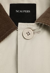 Camisa de tela beige con un cuello de pana marrón oscuro, con cierre de botones y una etiqueta negra con el nombre de la marca "SCALPERS."