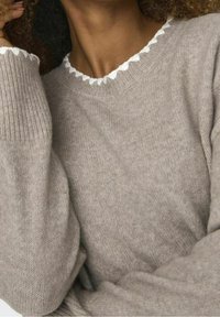 Hellbraunes Strickpullover mit gerippter Textur, ausgestattet mit einem gewellten weißen Saum am Ausschnitt und lässig geschnittenen Ärmeln.