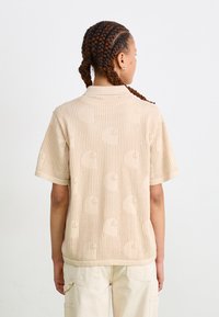 Short-sleeve polo shirt in beige with a textured knit and repeating logo pattern. Features a collar and ribbed hem. Relaxed fit.