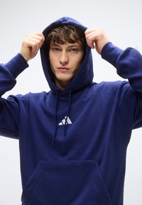 Tengerészkék kapucnis pulóver elején zsebbel, húzózsinóros kapucnival és a mellrészen fehér háromszög alakú logóval. Puha anyag, sima textúrával.