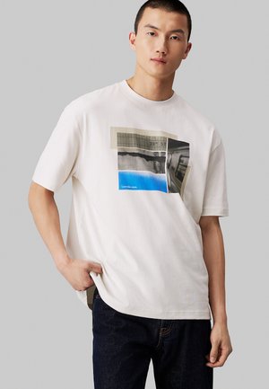 Camiseta de algodón blanca con estampado gráfico en tonos de gris y azul, que presenta patrones rectangulares y detalle del logo, corte relajado y cuello redondo.