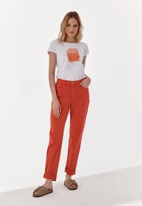 White t-shirt with a coral paint stroke design, paired with rolled-up, bright coral trousers. Light brown slide sandals complete the look.