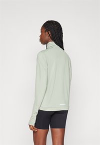 Camiseta atlética de manga larga en verde claro con un alto cuello y agujeros para los pulgares, combinada con pantalones cortos negros. Textura suave y acentos de diseño mínimos.