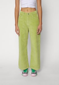 Pantalones de pana de pierna ancha en verde brillante. Tejido texturizado con estrías verticales. Llevados con zapatillas verdes y rosas. Bolsillos frontales visibles.