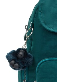 Mochila azul verdoso con un llavero de gorila peludo azul marino enganchado cerca de un bolsillo con cremallera sobre un fondo blanco.