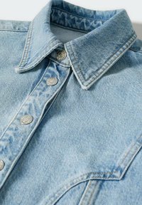 Veste en jean bleu clair avec boutons-pression argentés, col pointu et surpiqûres détaillées sur le col et la poche poitrine.