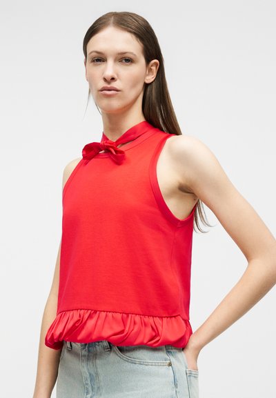 Γυναίκα που φοράει αμάνικο κόκκινο τοπ με φιόγκο στο ντεκολτέ και ραβδώσεις στο τελείωμα, συνδυασμένο με ανοιχτόχρωμο μπλε τζιν, πάνω σε απλό ανοιχτό φόντο.