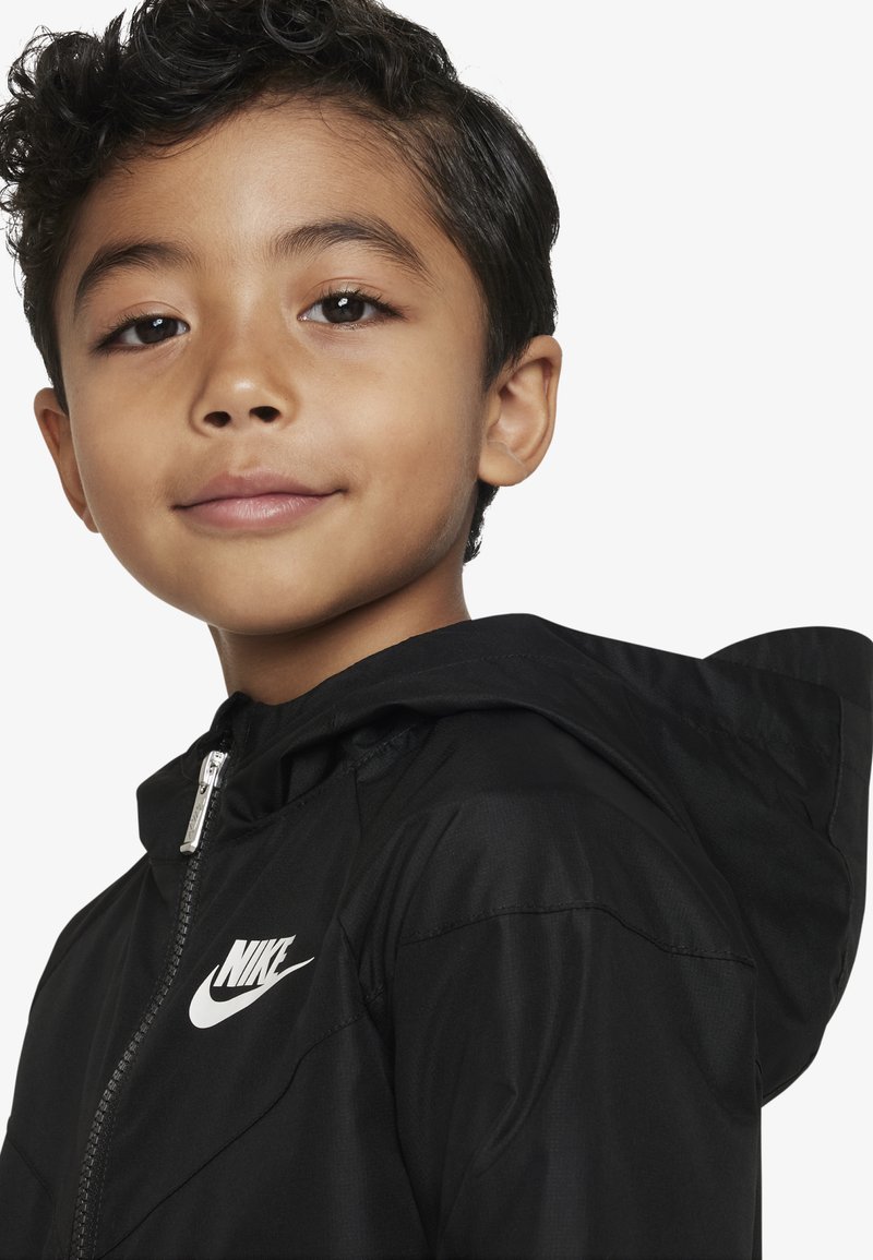 Czarna kurtka z kapturem Nike z zamkiem z przodu, wykonana z lekkiego materiału i wyróżniającego się białego logo na klatce piersiowej. Gładka tekstura.