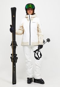 Skidragt bestående af en hvid og beige jakke, hvide bukser, sorte skistøvler, sorte ski med guldaccenter og sorte handsker.