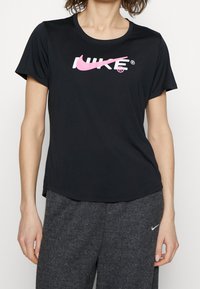 Camiseta Nike de manga corta negra con un logo rosa en el pecho. Cuenta con un cuello redondo y un corte holgado.