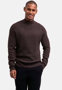 Brauner gestrickter Pullover mit hohem Kragen, geripptem Muster und langen Ärmeln, kombiniert mit einer dunklen Hose. Die Passform ist körpernah.