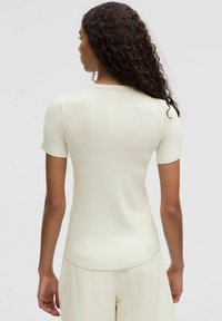 Priliehavý top s krátkym rukávom v svetlej krémovej pruhovanej látke. Má okrúhly výstrih a jemný stredový šev na zadnej strane. Hladká textúra.