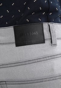 Pantaloni in denim grigio chiaro con etichetta in pelle nera con testo in rilievo. Il materiale ha una texture liscia con dettagli di cucitura sottili.