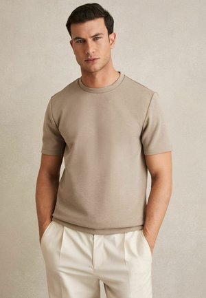 Tricou bej cu mânecă scurtă, confecționat dintr-un material neted, având un gât rotund și o croială relaxată. Asortat cu pantaloni plisati de culoare deschisă.