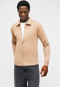 Άνδρας με μπεζ ζακέτα με φερμουάρ πάνω από λευκό πουκάμισο και σκούρα γκρι τζιν, στέκεται μπροστά σε απλό ανοιχτό φόντο.