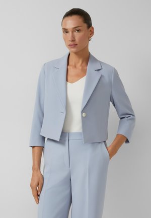 Vrouw draagt een lichtblauwe cropped blazer met een enkele knoop, bijpassende broek en een witte V-hals top tegen een effen grijze achtergrond.