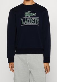 Sudadera navy con cuello redondo ribeteado y puños. Cuenta con un logo de cocodrilo verde y el texto "LACOSTE" en blanco arriba. Tejido suave.