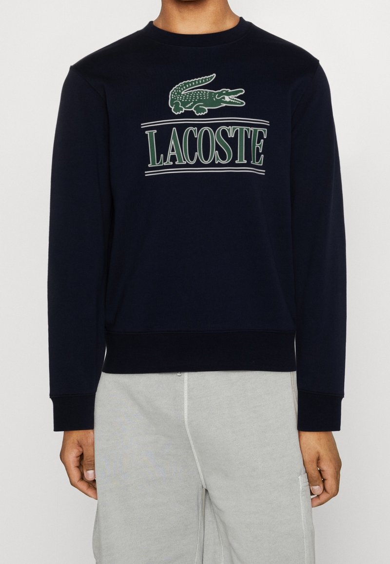 Sudadera navy con cuello redondo ribeteado y puños. Cuenta con un logo de cocodrilo verde y el texto "LACOSTE" en blanco arriba. Tejido suave.