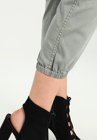 Grijze cropped broek met elastische boorden, gecombineerd met zwarte enkellaarzen met uitsnijdingen en veters. Glad stofoppervlak zichtbaar.