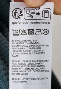 Hoito-ohjeiden etiketti kevyessä kankaassa, jossa on huolto-ohjeet. Teksti sisältää materiaalit: 93% polyesteria, 7% spandeksi; 95% polyesteria, 5% elastaania.