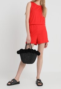 Czerwony kombinezon bez rękawów z dopasowaną talią, czarna pleciona torebka koszykowa ozdobiona czarnymi pomponami oraz czarne klapki z ćwiekami.