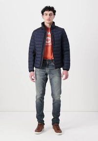 Jachetă puffer navy cu design matlasat, tricou portocalie, blugi albaștri și adidași maronii. Jacheta are un guler înalt și închidere cu fermoar.