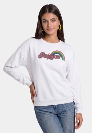 Giovane donna con lunghi capelli scuri che indossa orecchini a cerchio gialli, una felpa bianca con testo "Rainbow" e grafica arcobaleno, e jeans blu.