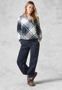Camicia a quadri con sfumature di blu navy, bianco e turchese; pantaloni in denim a vestibilità ampia; stivali marroni con lacci; in piedi contro uno sfondo grigio texturizzato.