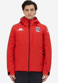 Giacca isolante rossa con zip frontale, cappuccio e logo Kappa. Presenta toppe della US Ski Team, polsini elasticizzati e tasche laterali.