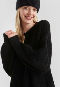 Jonge vrouw die glimlacht, draagt een zwarte gebreide beanie en een oversized zwarte pluizige trui, tegen een effen lichtgrijze achtergrond.